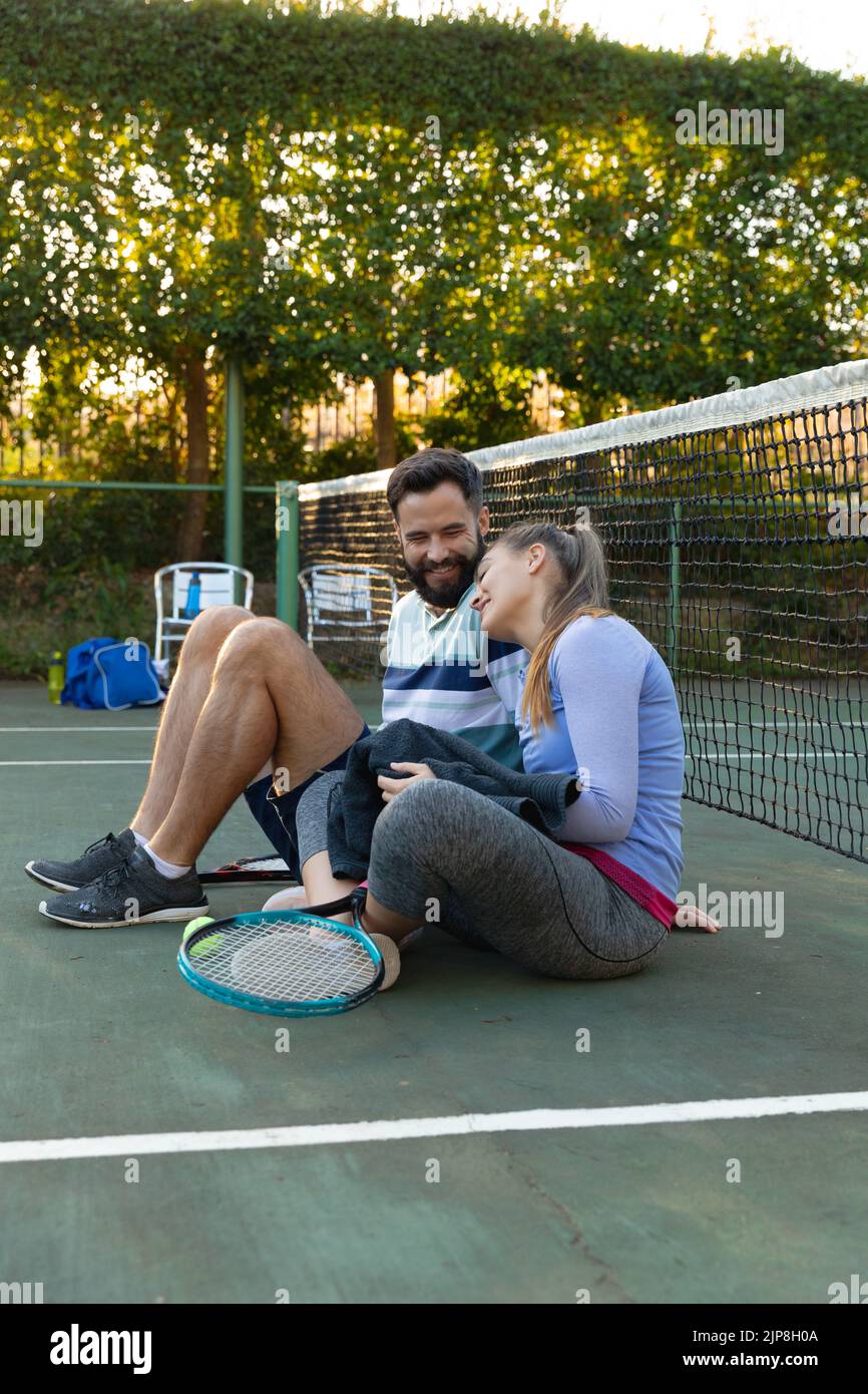 Glückliches kaukasisches Paar, das auf dem Tennisplatz im Freien im Netz sitzt und sich nach dem Tennisspielen erholt Stockfoto