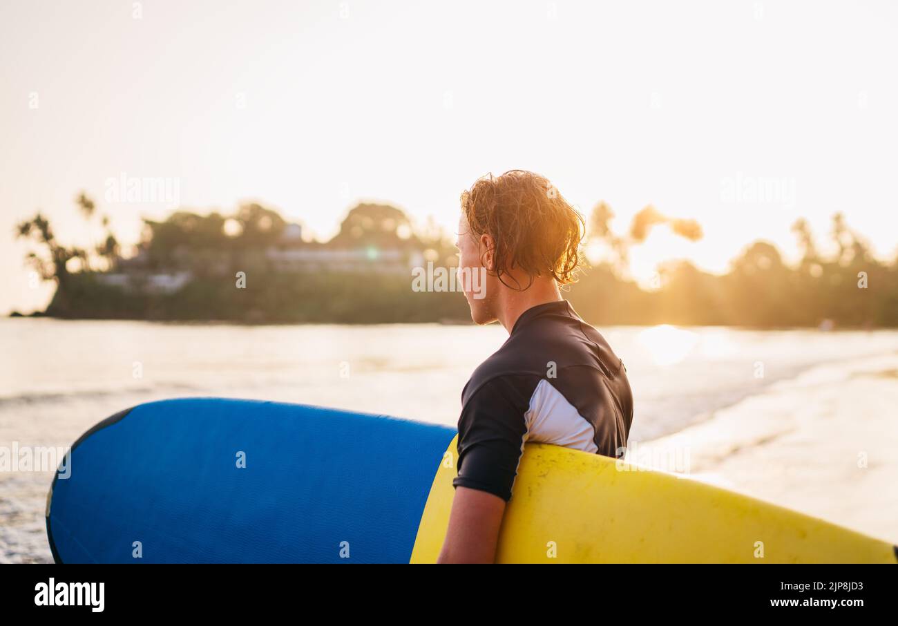 Junger Teenager Junge mit buntem Surfbrett geht zum Surfen ins Meer. Er genießt ein wunderschönes Sonnenuntergangslicht am Dewata Strand auf Sri Lanka. Aktiver Sport Stockfoto