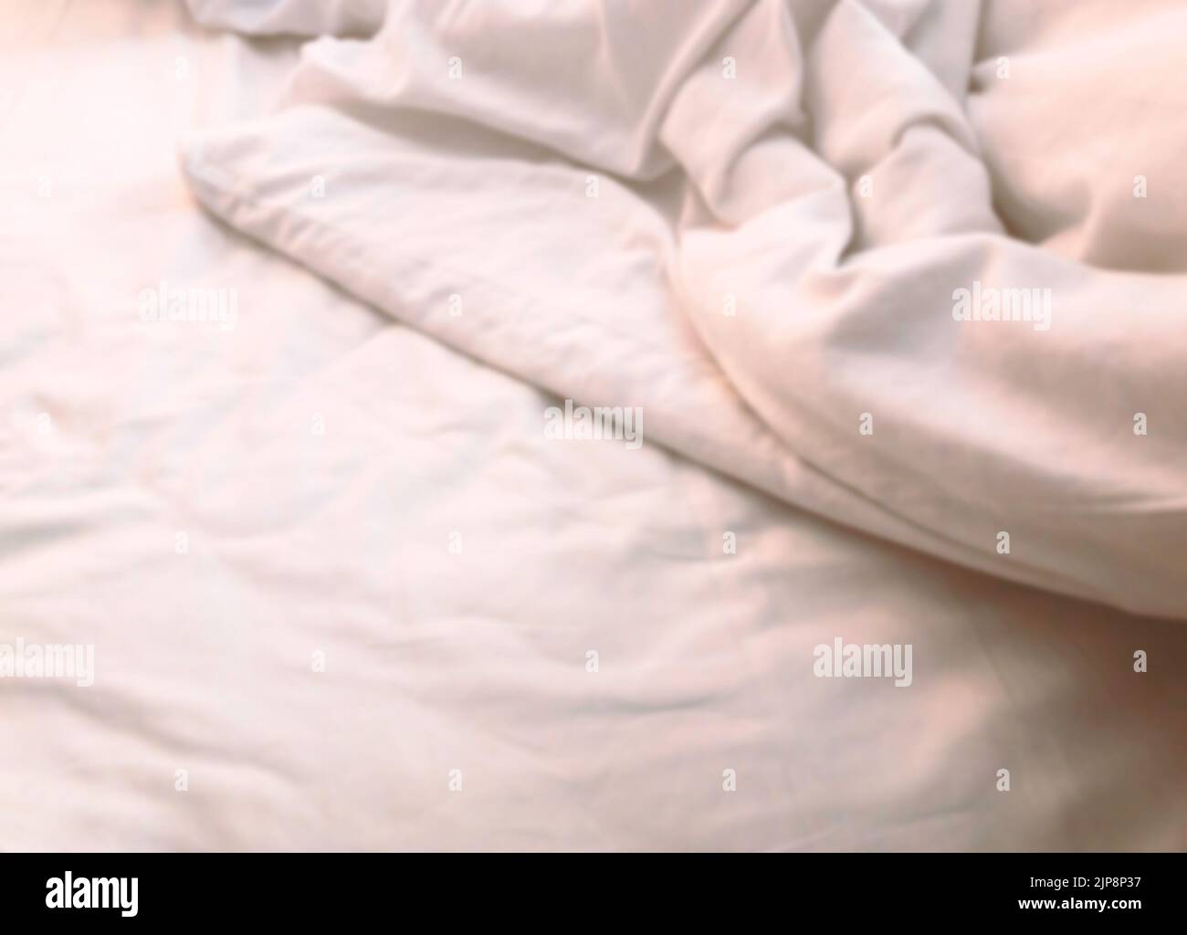 Verschwommenes Bild ein Teil des weißen Bettes mit Decke, Nahaufnahme des leeren Raumes auf dem weißen Bett, Falten auf dem Bett, warmes Licht. Stockfoto