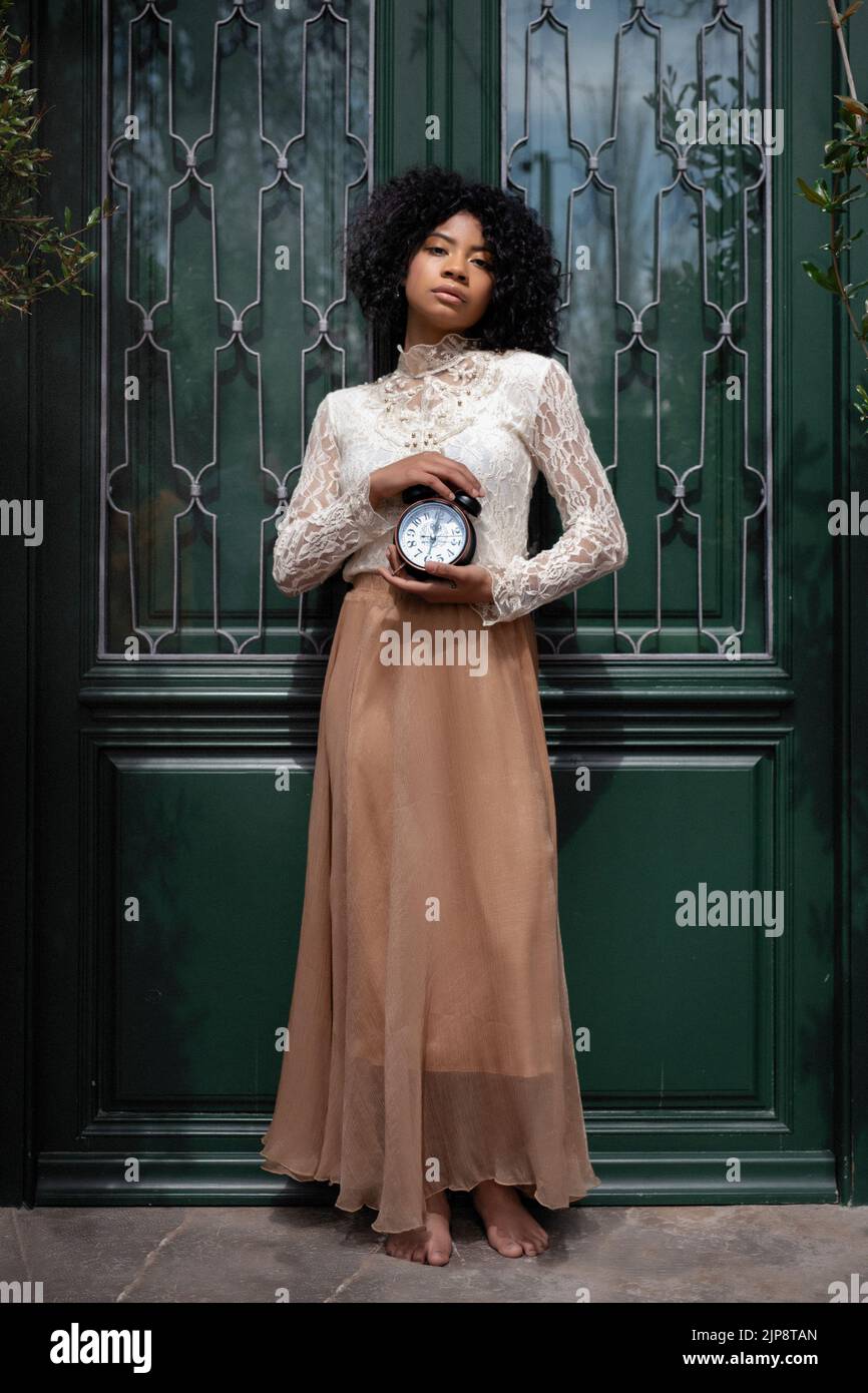 Voller Körper von ruhigen jungen barfüßigen ethnischen Frauen mit afrodunklem Haar im Retro-Stil Outfit, das neben grünen Türen mit Vintage-Wecker und Look steht Stockfoto