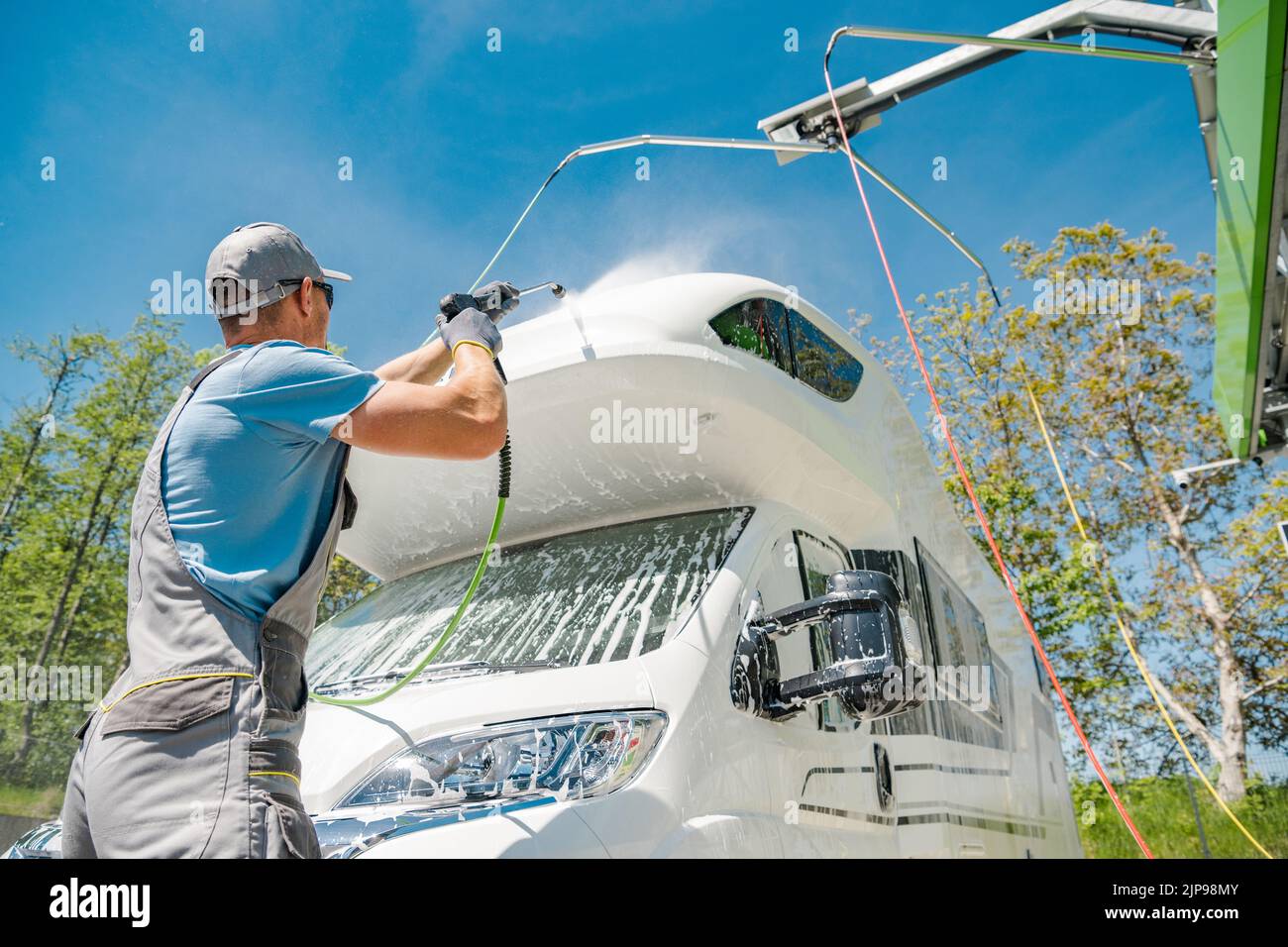 Rückansicht des kaukasischen männlichen Drucks beim Waschen seines weißen Wohnmobils bei der lokalen Autowäsche. Sonniger Sommertag mit sauberem blauen Himmel im Hintergrund. Stockfoto