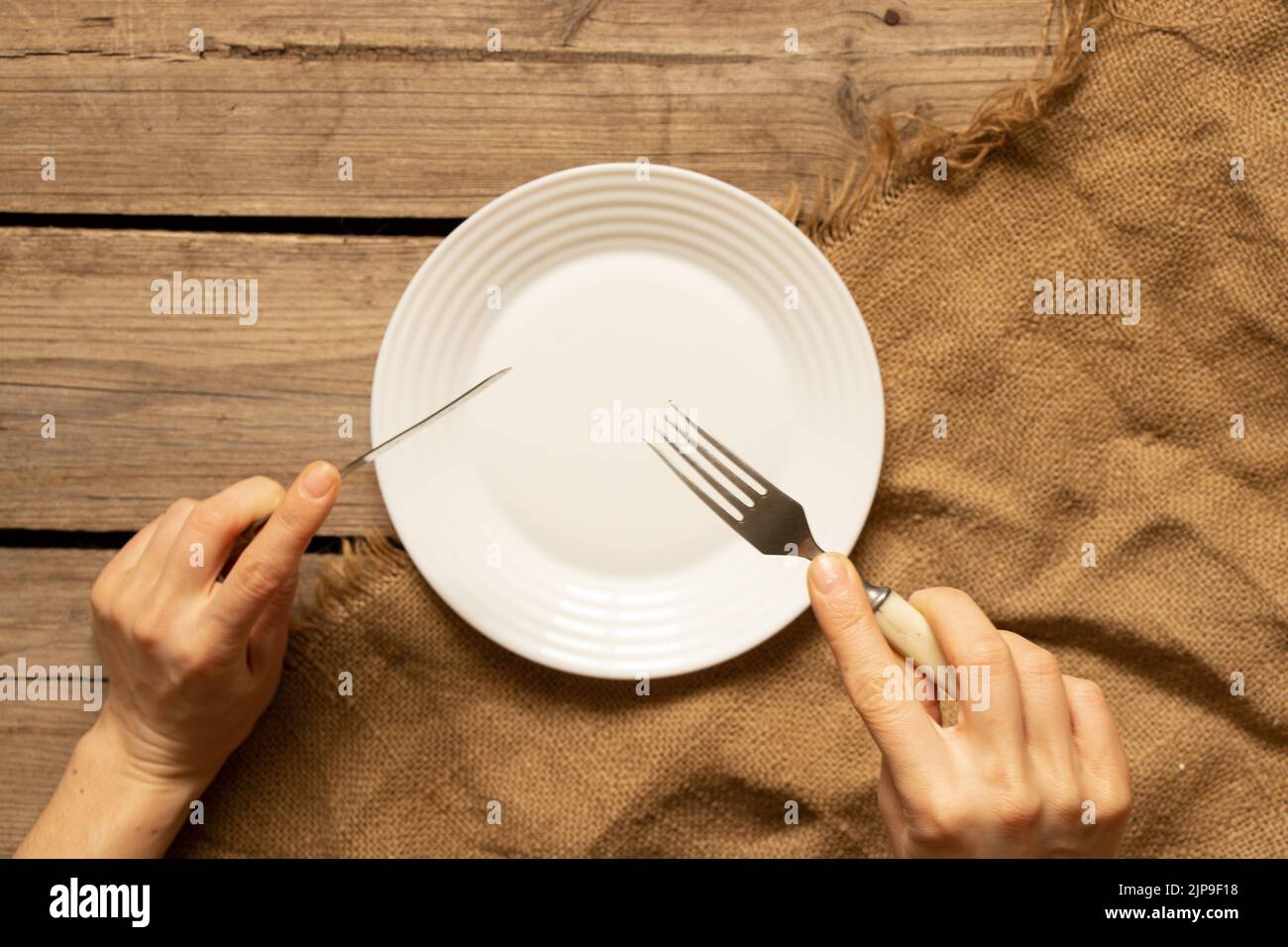 Ein weißer Teller und eine Gabel mit einem Messer in den Händen auf einem alten Holztisch in der Küche zu Hause, Restaurant Business, Küchengeräte Stockfoto