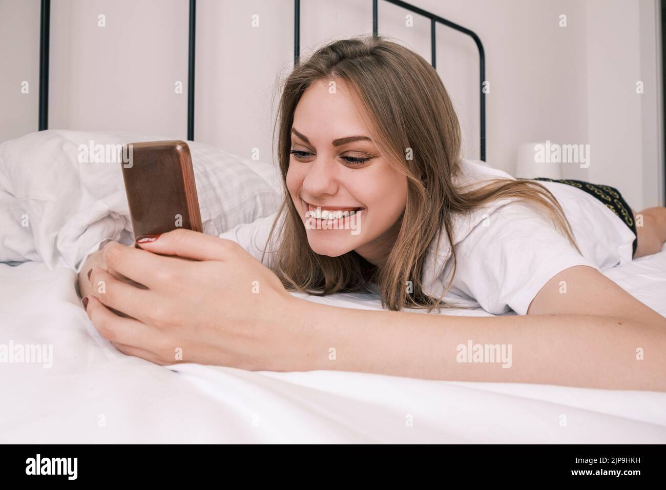 Junge, fröhlich lächelnde Frau, die morgens im Bett lag und ihr Telefon benutzte. Erwachen. Mädchen liest gute Nachrichten, soziale Netzwerke, Online-Kommunikation Stockfoto