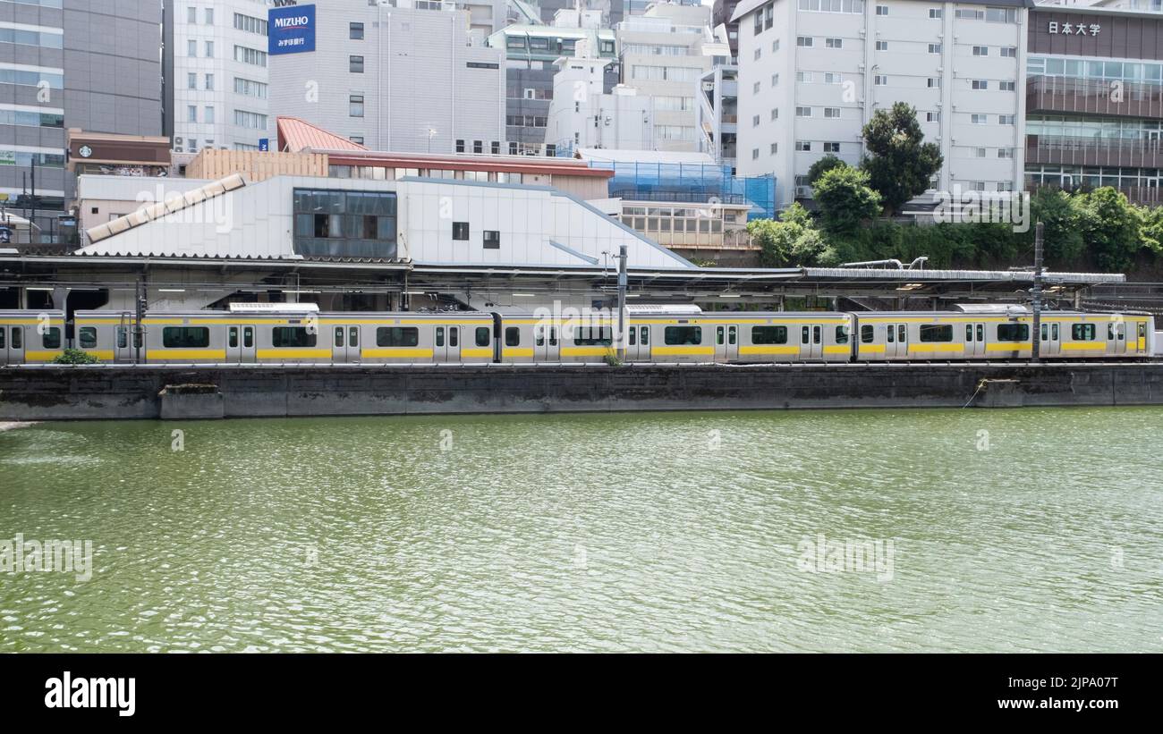 Der JR-Bahnhof Ichigaya liegt an einem Fluss mit dem gelben Zug der U-Bahn-Shinjuku-Linie Toei an einem bewölkten Sommertag. Stockfoto