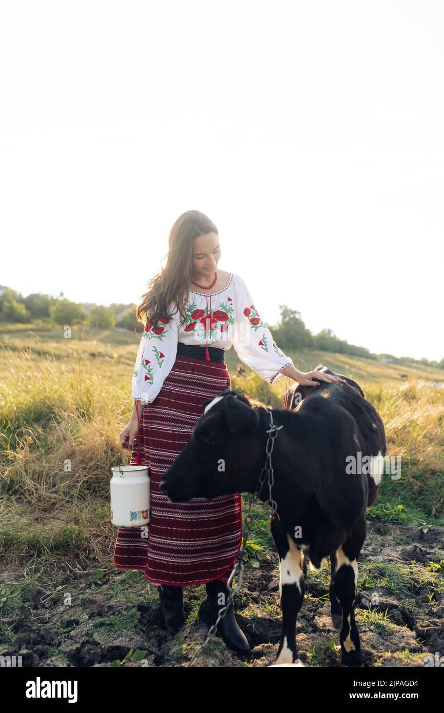 Junge ukrainische Frau steht in der Nähe von Kalb mit Wasserbüchse in traditionellen nationalen bestickten Hemd und Rock auf der Weide gekleidet. Ethnische ukrainische nationa Stockfoto