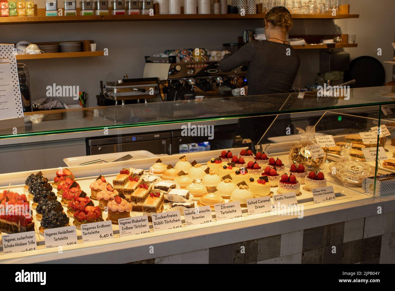 Ausgefallene schöne Desserts werden in einer Glasvitrine mit einem Hintergrund einer Frau angezeigt, die Kaffee zubereitet Stockfoto