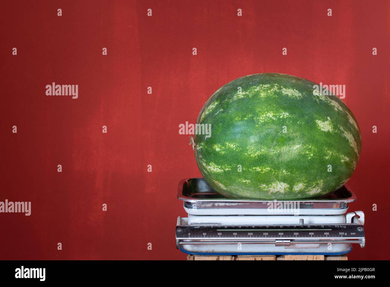 Eine alte Küchenwaage steht vor einem roten Hintergrund mit Platz für Text. Auf der Waage befindet sich eine große grüne Wassermelone Stockfoto