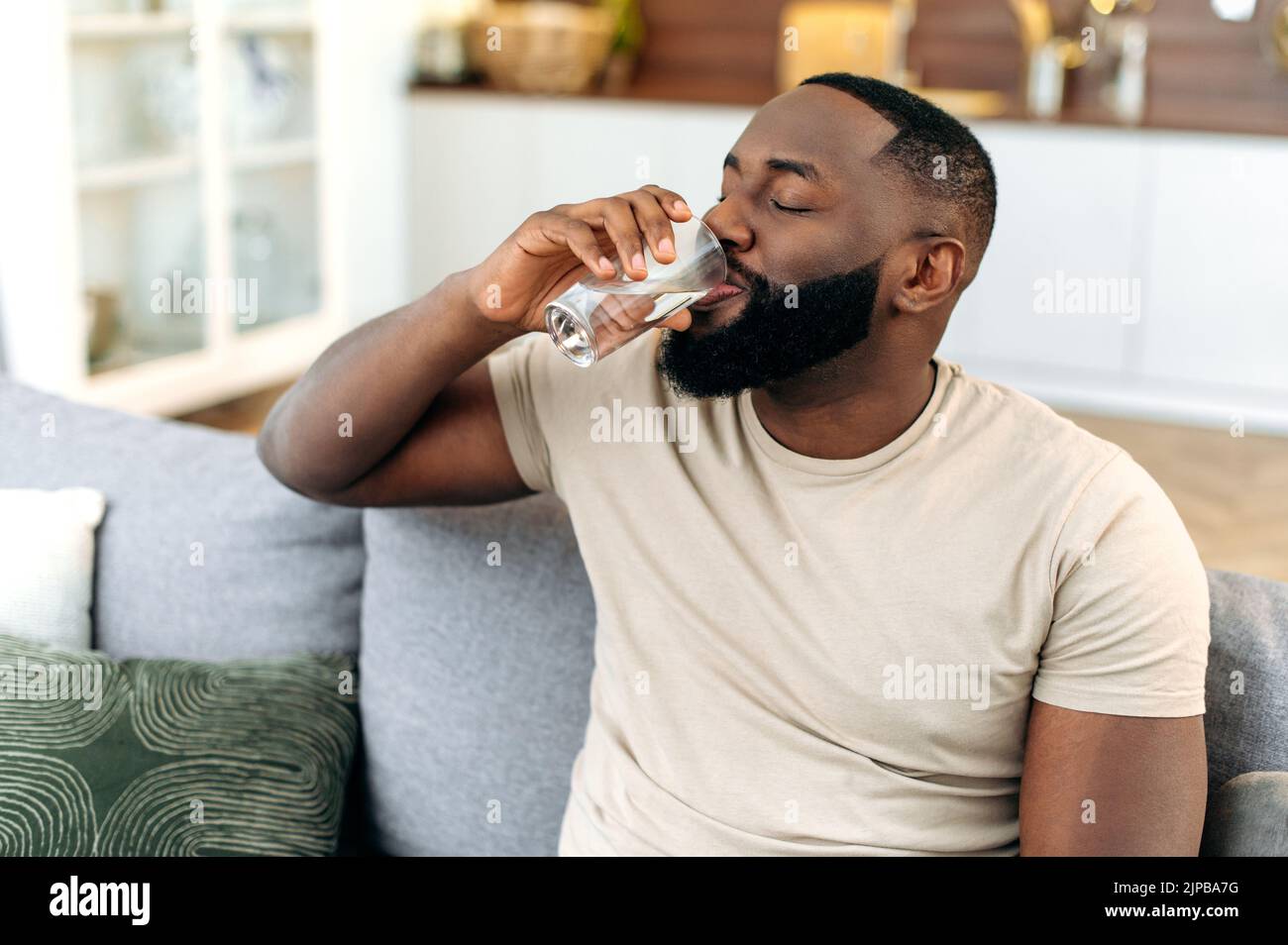 Gesunder Lebensstil. Ein positiver afroamerikanischer Mann, sitzt zu Hause auf dem Sofa und trinkt ein Glas sauberes Wasser. Schöner schwarzer Mann führt einen gesunden Lebensstil, überwacht den Wasserhaushalt im Körper Stockfoto