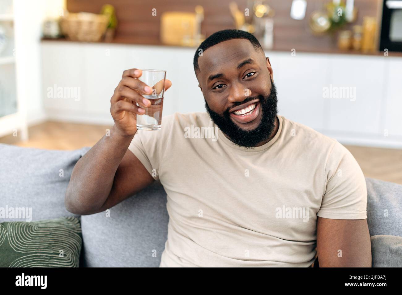 Glücklicher afroamerikanischer Kerl, sitzt zu Hause auf dem Sofa, hält ein Glas sauberes Wasser in der Hand, blickt auf die Kamera und lächelt. Schöner schwarzer Kerl führt einen gesunden Lebensstil, überwacht den Wasserhaushalt im Körper Stockfoto