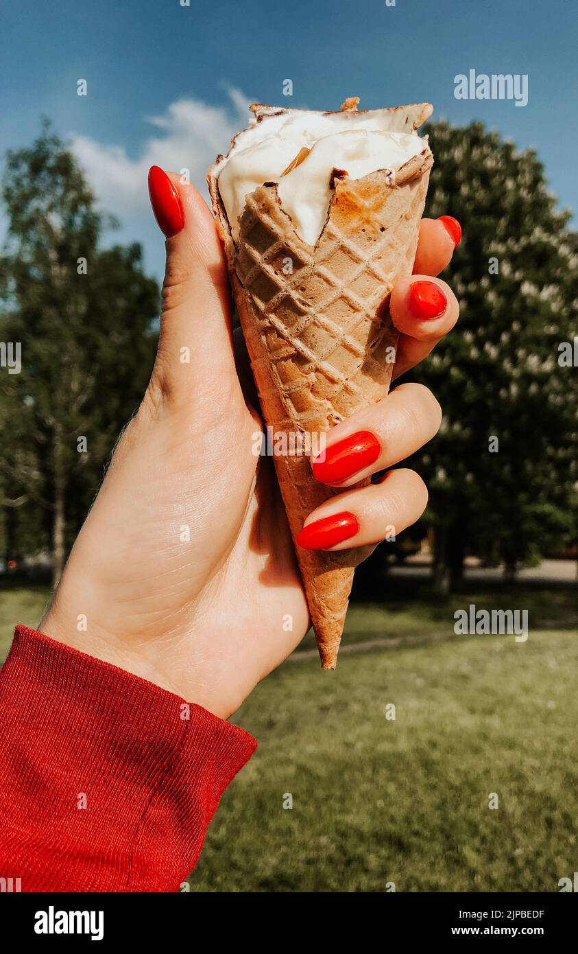 Frau Hand hält Eis am sonnigen Sommertag Nahaufnahme. Natürliche grüne Hintergrund. Rote Maniküre Mädchen Nägel. Eis-Kegel in einem Waffelbecher. Sommer Stockfoto