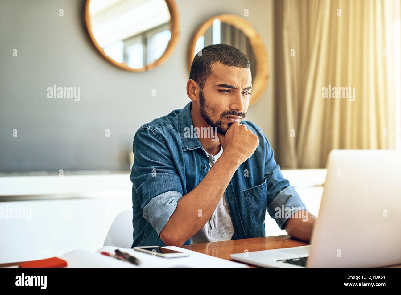 Ich muss hart darüber nachdenken: Ein fokussierter junger Mann, der an seinem Laptop arbeitet, während er zu Hause hinter einem Tisch nachdenkt. Stockfoto