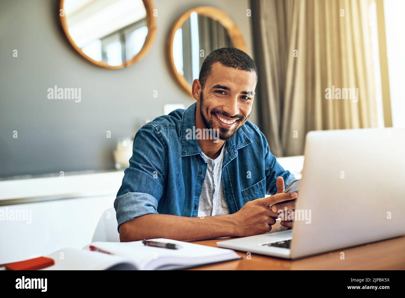 Ich machte heute von zu Hause aus große Fortschritte. Porträt eines fröhlichen jungen Mannes, der an seinem Laptop arbeitet und sein Telefon überprüft, während er an einem Tisch sitzt Stockfoto