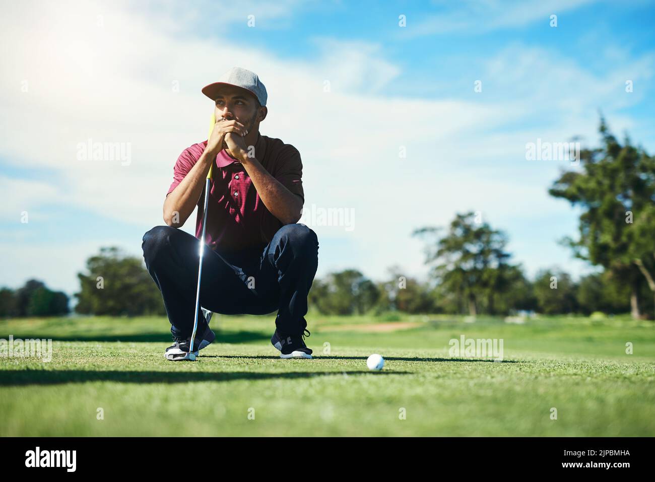 Auf dem Fairway. Ein fokussierter junger Golfer, der einen Golfball anschaut, während er tagsüber draußen auf dem Gras sitzt. Stockfoto