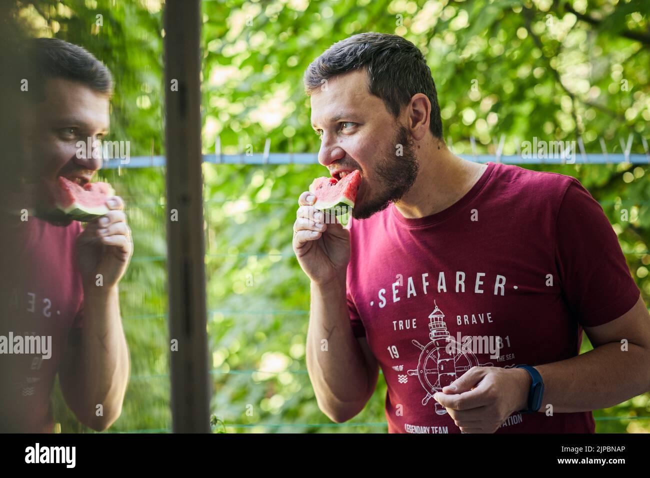 Mann, der saftige rote Wassermelone auf dem Balkon isst. Das Konzept von Sommer und köstlichem Gemüse. Vorderansicht. Stockfoto