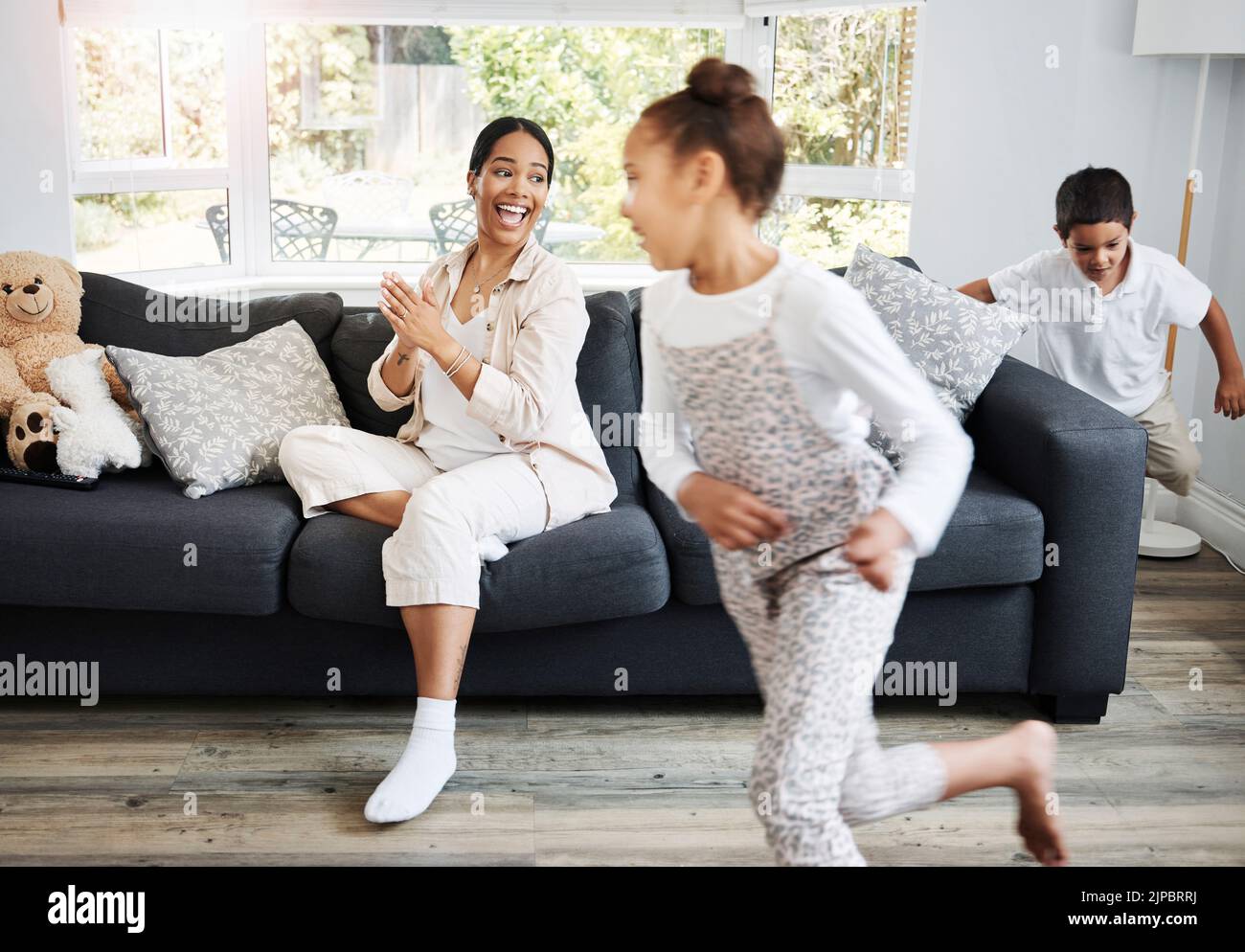 Spielen, Laufen und kleine Kinder aufgeregt drinnen mit einer lächelnden Mutter beobachten auf einer Couch. Glückliche Familie, die den Tag zusammen drinnen verbringt Stockfoto