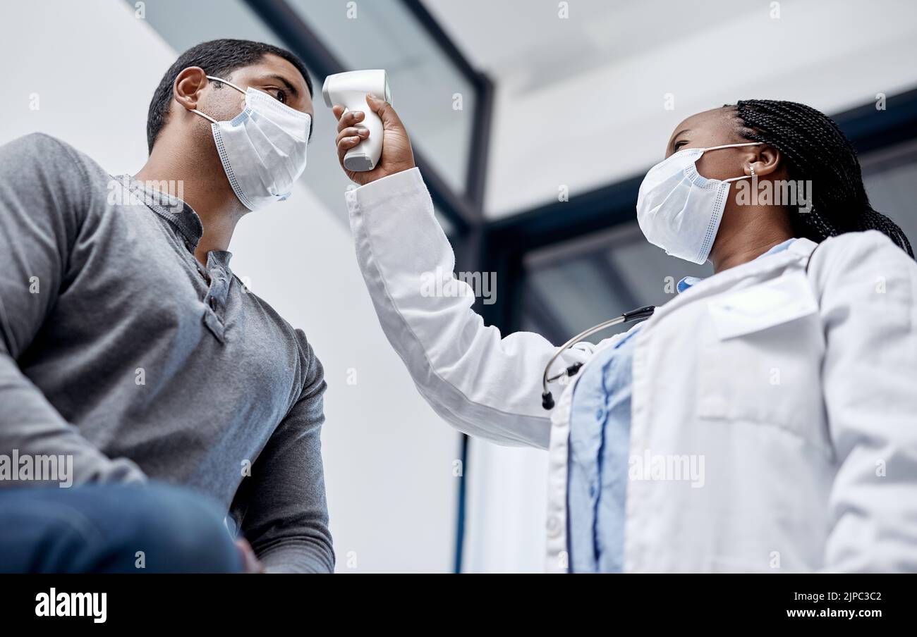 Ärztin, die einen männlichen Patienten mit Augenwippung konsultiert und seine Temperatur mit einem Infrarot-Thermometer im Krankenhauszimmer oder in der Klinik einnimmt. Mann trägt Maske und Stockfoto