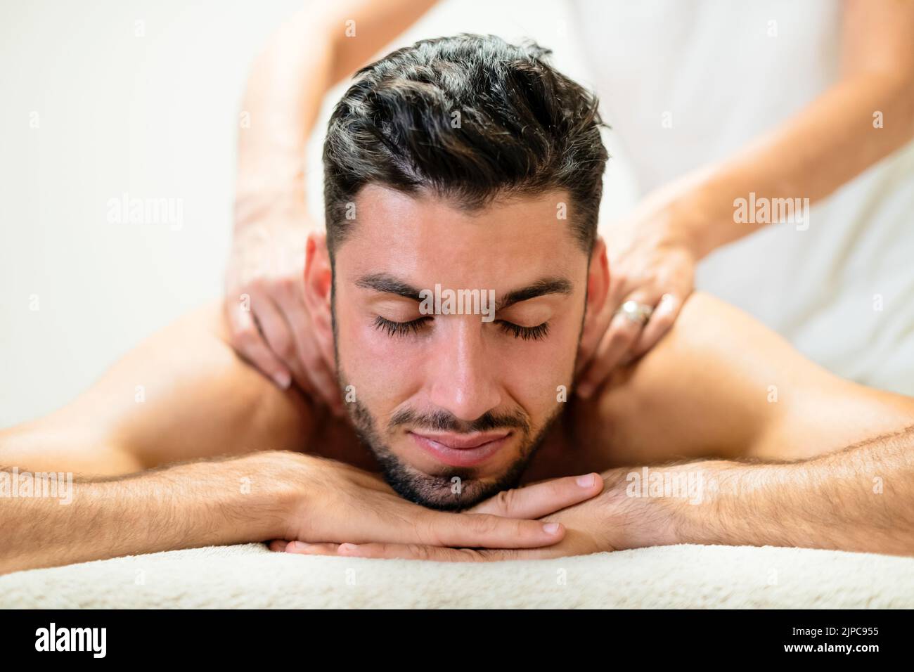 Ruhiger junger Mann, der die Arme unter das Kinn faltete und die Augen schloss, während er während der Schultermassage im Spa-Salon auf dem Tisch lag Stockfoto