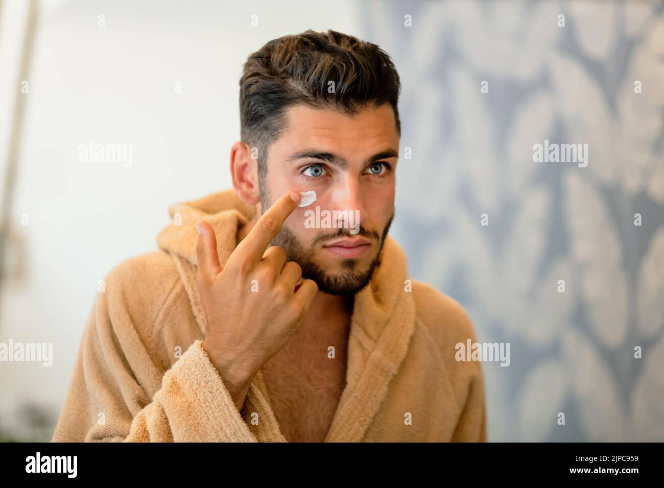 Junger bärtiger Mann im Bademantel, der während der morgendlichen Hautpflege auf den Spiegel schaut und feuchtigkeitsspendende Creme unter blauem Auge schmiert Stockfoto