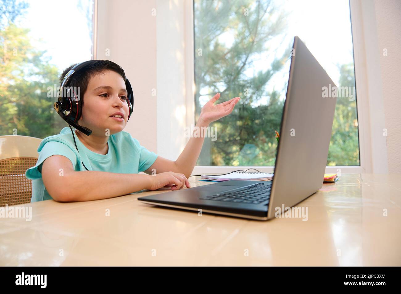 Intelligenter Schuljunge mit kabellosen Kopfhörern sieht sich Online-Videostunden an, kommuniziert mit dem Lehrer, studiert von zu Hause aus Stockfoto