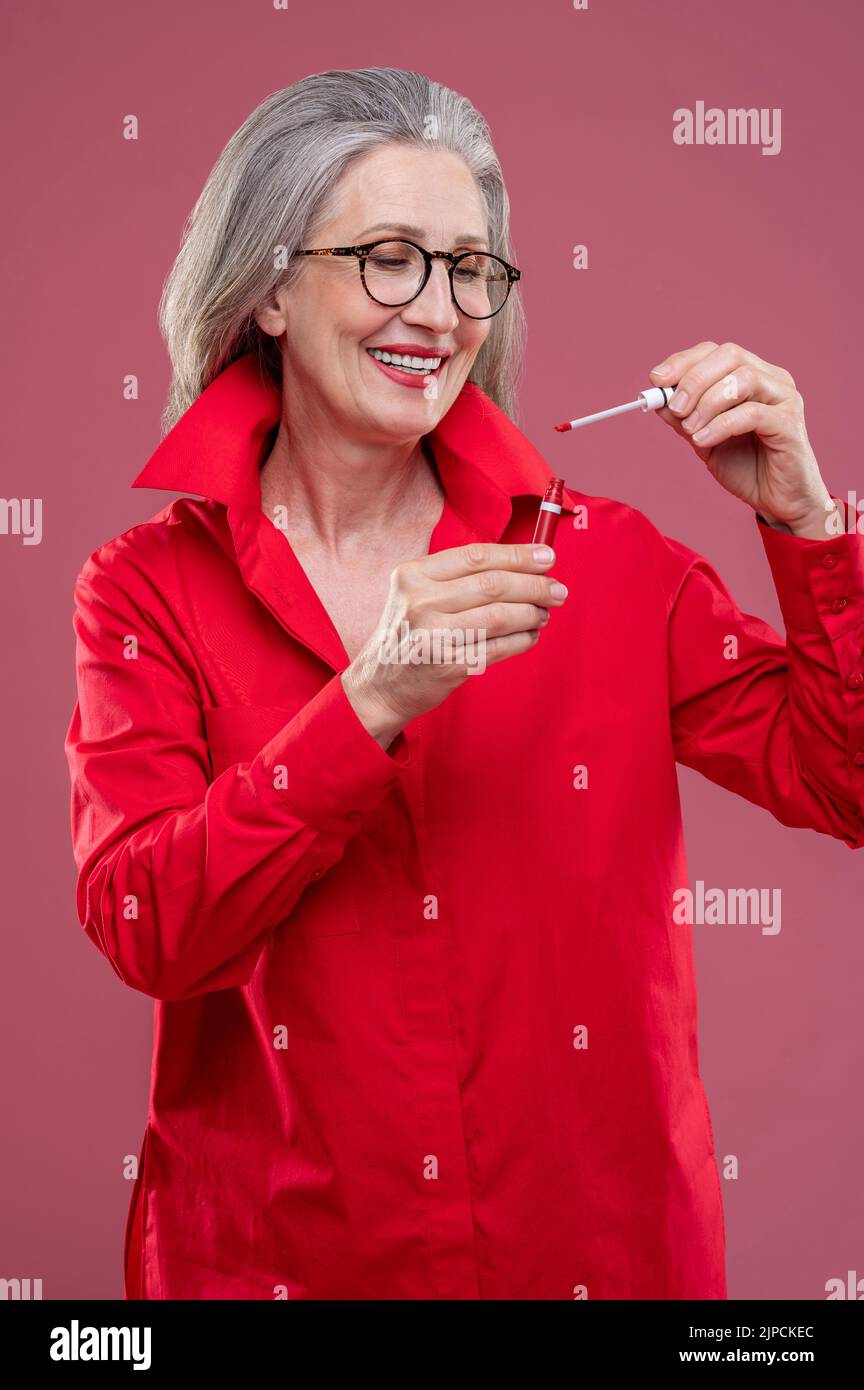 Frau in rot leuchtend Hemd hält Lipgloss und sieht zufrieden Stockfoto