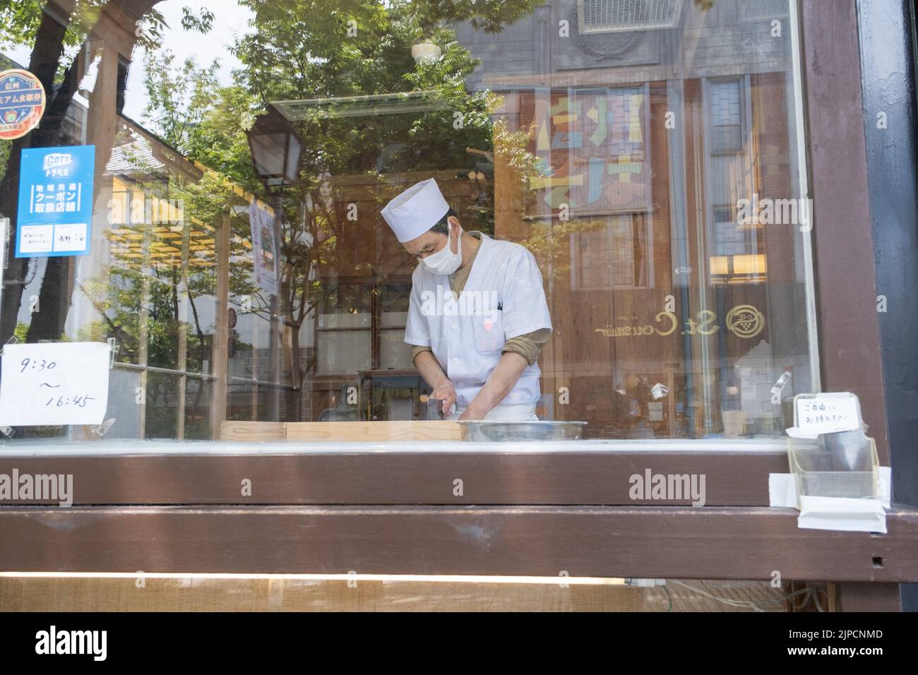 Ein Schuss durch das Fenster eines Soba-Kochs im Zenko-Ji-Tempel im Sommer, der eine Maske und einen Hut trägt und handgemachte Buchweizen-Nudeln zum Mittagessen zubereitet. Stockfoto