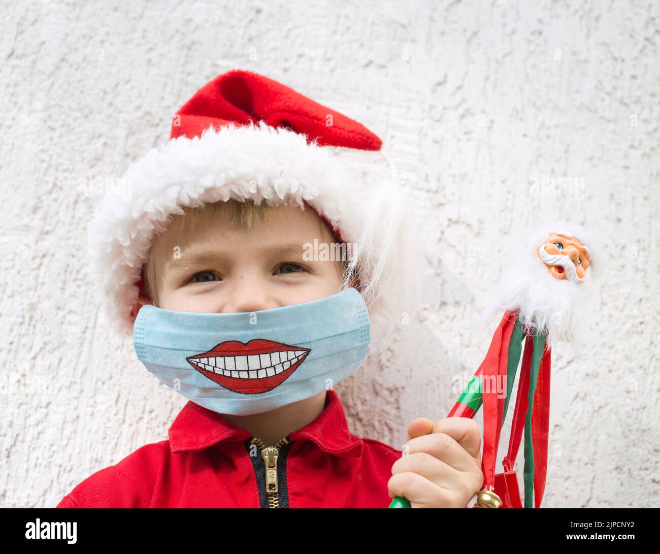 Kind in Santa Hut . Auf dem Gesicht befindet sich eine blaue medizinische Maske mit gemaltem, witzigem Lächeln. Zeit für Geschenke. Bleib zu Hause. Tragen Sie Masken. Positives Denken. Christus Stockfoto