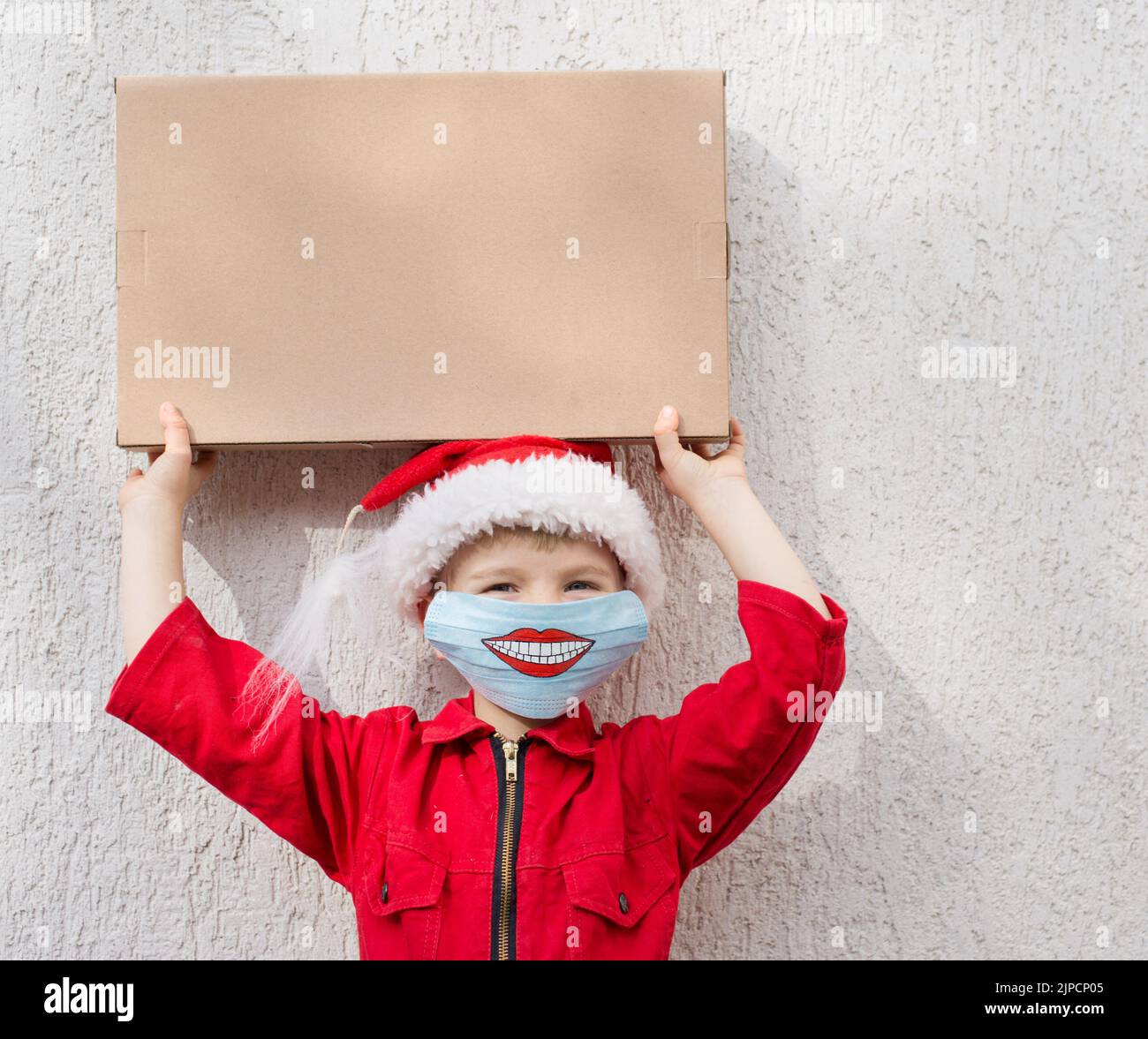 Neukäufe oder Lieferung aus dem Geschäft. Kleinkind Junge in roten Jumpsuit, Weihnachtsmütze. Auf dem Gesicht ist blaue medizinische Maske mit Lächeln auf sie gemalt. Hält Pappe Stockfoto