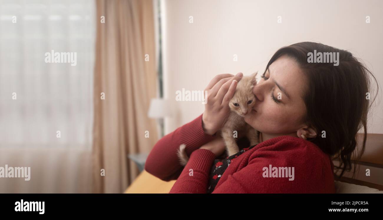 Portrait der schönen hispanischen jungen Frau mit roter Jacke umarmt und küsst ihr niedliches Haustier Baby Kätzchen in ihrem Zimmer mit unfokussierten Hintergrund Stockfoto