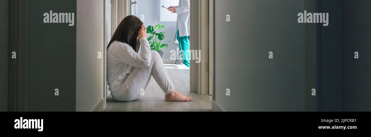 Patient, der auf dem Boden im Korridor eines psychiatrischen Zentrums sitzt, mit einem Arzt im Hintergrund Stockfoto