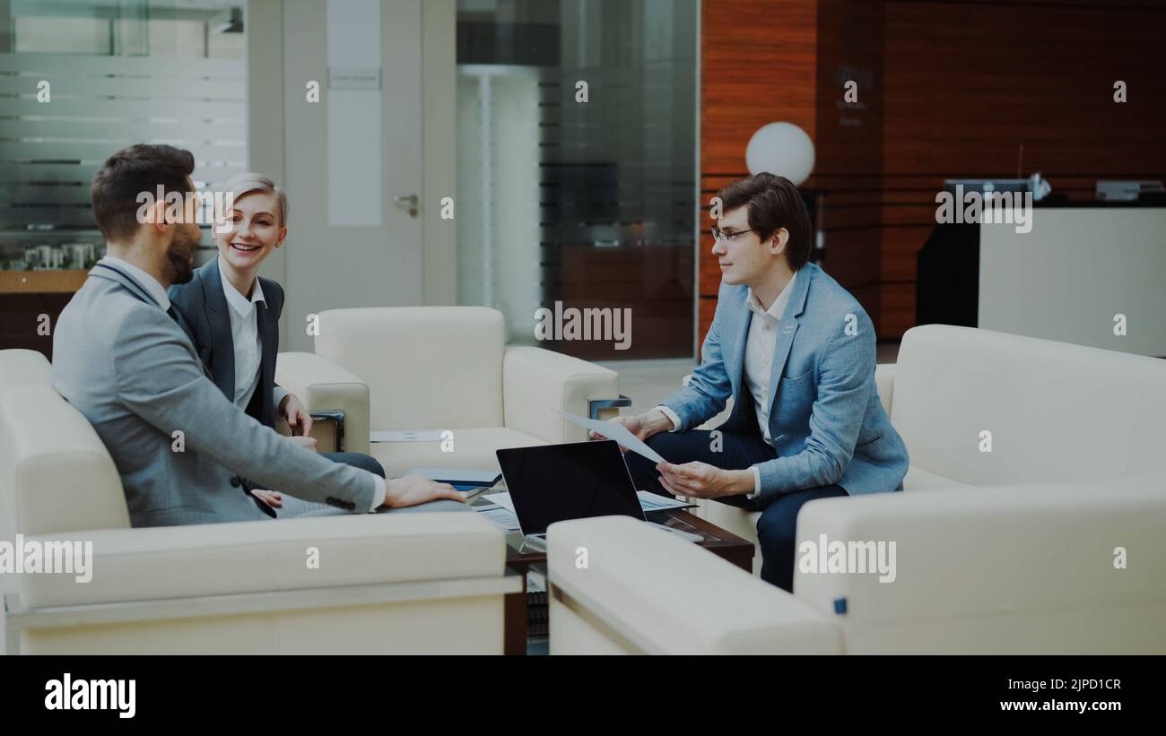 Fröhlicher Geschäftsmann, der Finanzberichte mit Geschäftspartnern diskutiert und auf dem Sofa in der modernen Bürohalle sitzt Stockfoto