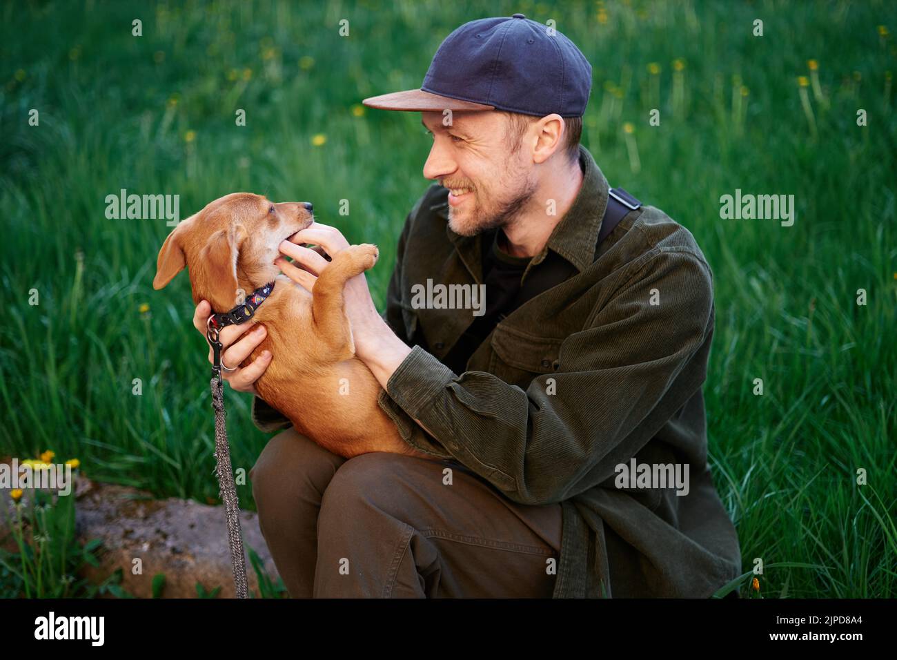 Hund als Familienmitglied Konzept. Attraktiver kaukasischer Mann in Baseballmütze, der auf einem Gras im Park sitzt und mit einem kleinen braunen Welpen spielt, der ihn am Finger beißt. Junger Mann mit Dackel im Freien Stockfoto
