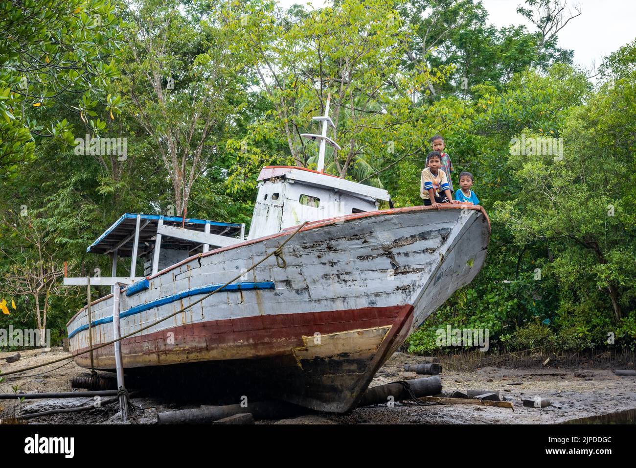 Drei kleine Jungen, die auf dem Bug eines verlassenen Fischerbootes sitzen. Sulawesi, Indonesien. Stockfoto