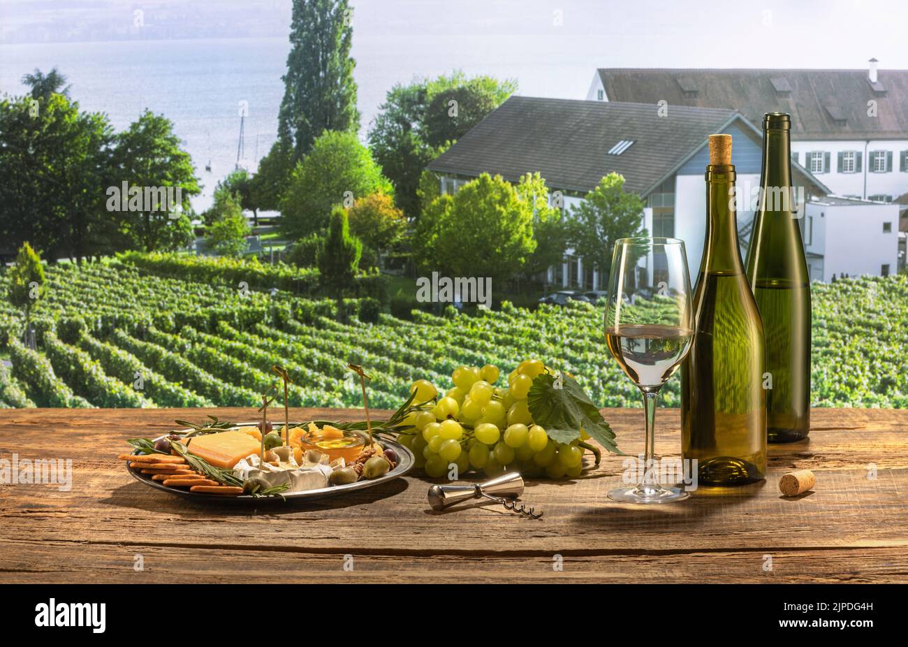 Zwei Flaschen und ein Glas köstlicher Weißwein stehen auf einem Holztisch. Leckere Vorspeisen mit Käse, Honig, Trauben und Crackern. Sommerweingarten Stockfoto