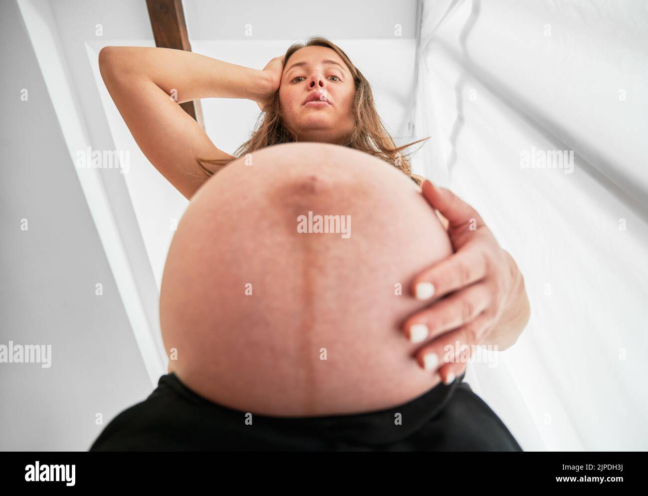 Bottom-Ansicht der Schwangeren, die durch die Größe ihres Bauches überrascht. Frau, die eine Hand auf ihren schwanger Bauch und die andere auf ihren Kopf legt und mit Bestürzung nach unten schaut. Stockfoto