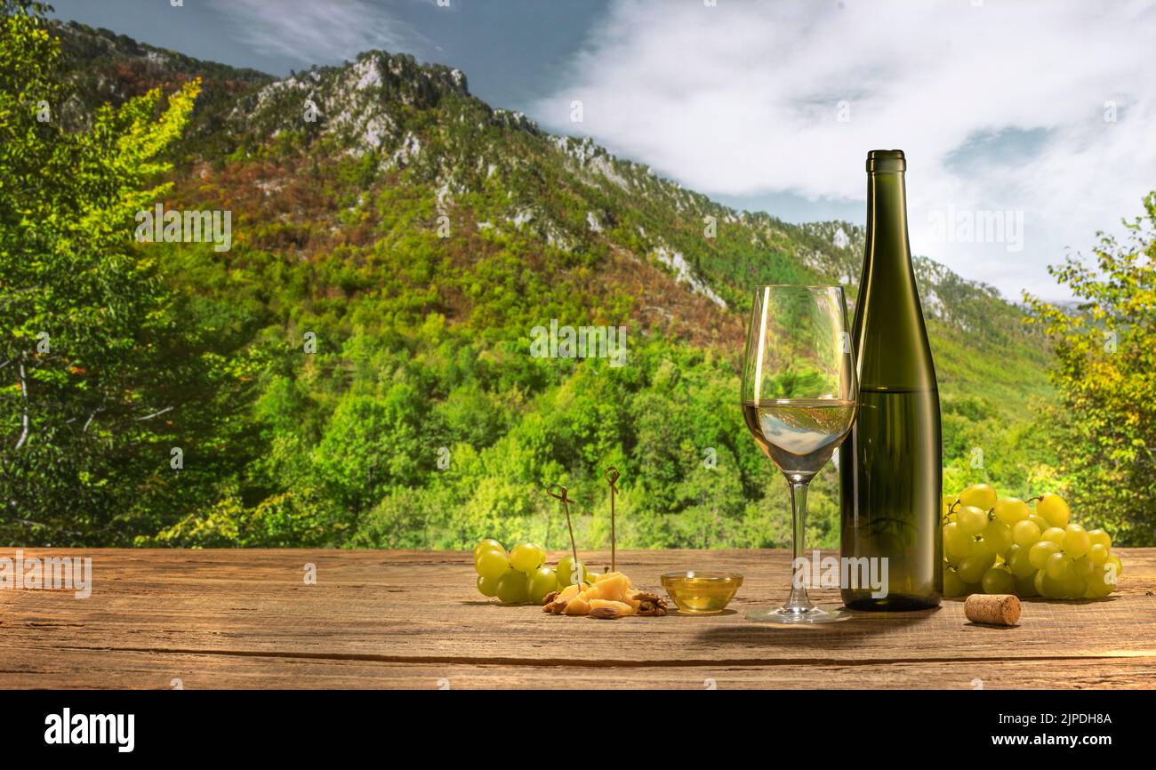 Besuch Weinberg im Sommer. Flasche und ein Glas Weißwein stehen auf einem Holztisch isoliert über dem Hintergrund der Berge am bewölkten Abend. Stockfoto