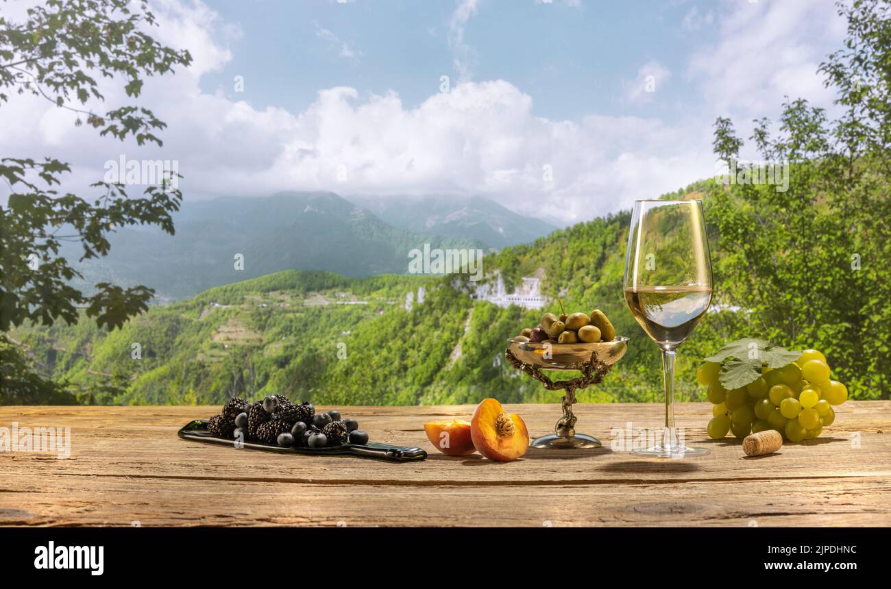 Oliven und Trauben. Flasche und ein Glas köstlicher Weißwein stehen auf einem Holztisch auf einer wunderschönen Naturlandschaft mit grünem Garten und Weinberg Stockfoto
