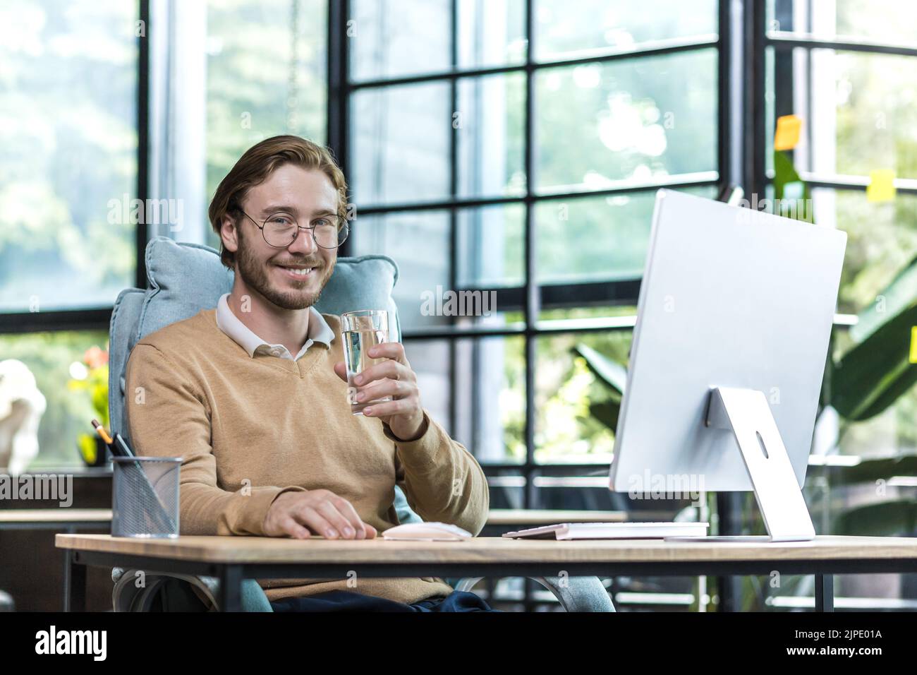 Junger erfolgreicher Geschäftsmann im modernen Büro bei der Arbeit lächelnd und mit Blick auf die Kamera, Geschäftsmann mit einem Glas Wasser, Arbeiter in Brillen und legerer Kleidung Stockfoto