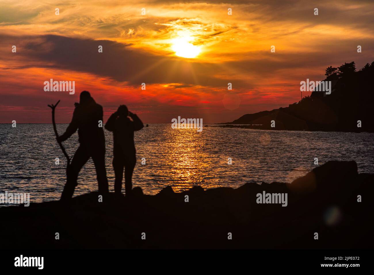 2 nicht identifizierbare Silhouetten von Menschen stehen auf Felsen mit Blick auf das Meer unter einer untergehenden Sonne. Einer von ihnen hält einen Stock. Stockfoto