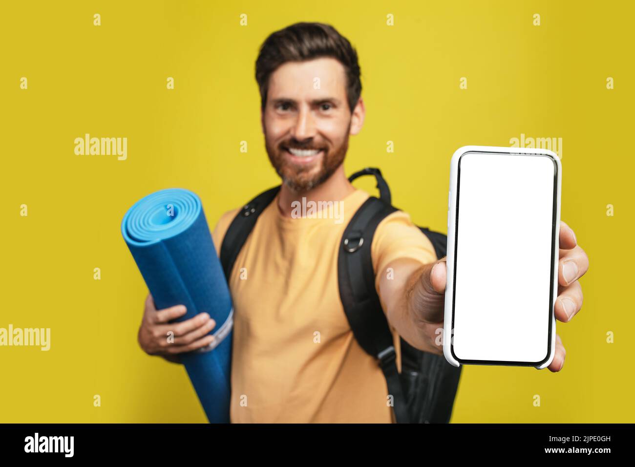 Online-Reisebüro. Aufgeregter männlicher Tourist, der sein Smartphone mit leerem Bildschirm zeigt und eine touristische Matte hält, Nachbau Stockfoto