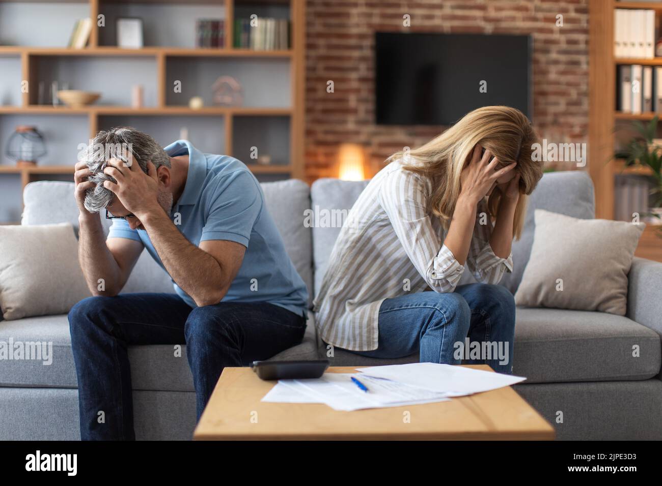 Unglückliches reifes kaukasisches Paar, das Köpfe mit Händen hält, leidet unter Schulden im Innenraum mit Dokumenten Stockfoto