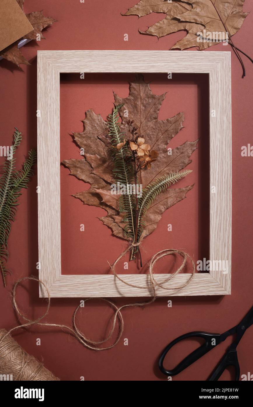 Bouquet aus getrocknetem gepresstem Blatt im Rahmen mit Schere, Seil und Notizbuch auf braunem Hintergrund. Hobby, handgemachte, florale Kunst und Boho-Stil Konzept. Stockfoto