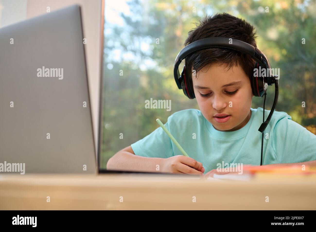 Nahaufnahme eines intelligenten, konzentrierten Schuljungen im Audio-Headset, der während des Online-Unterrichts mit einem externen Weblehrer Notizen macht Stockfoto