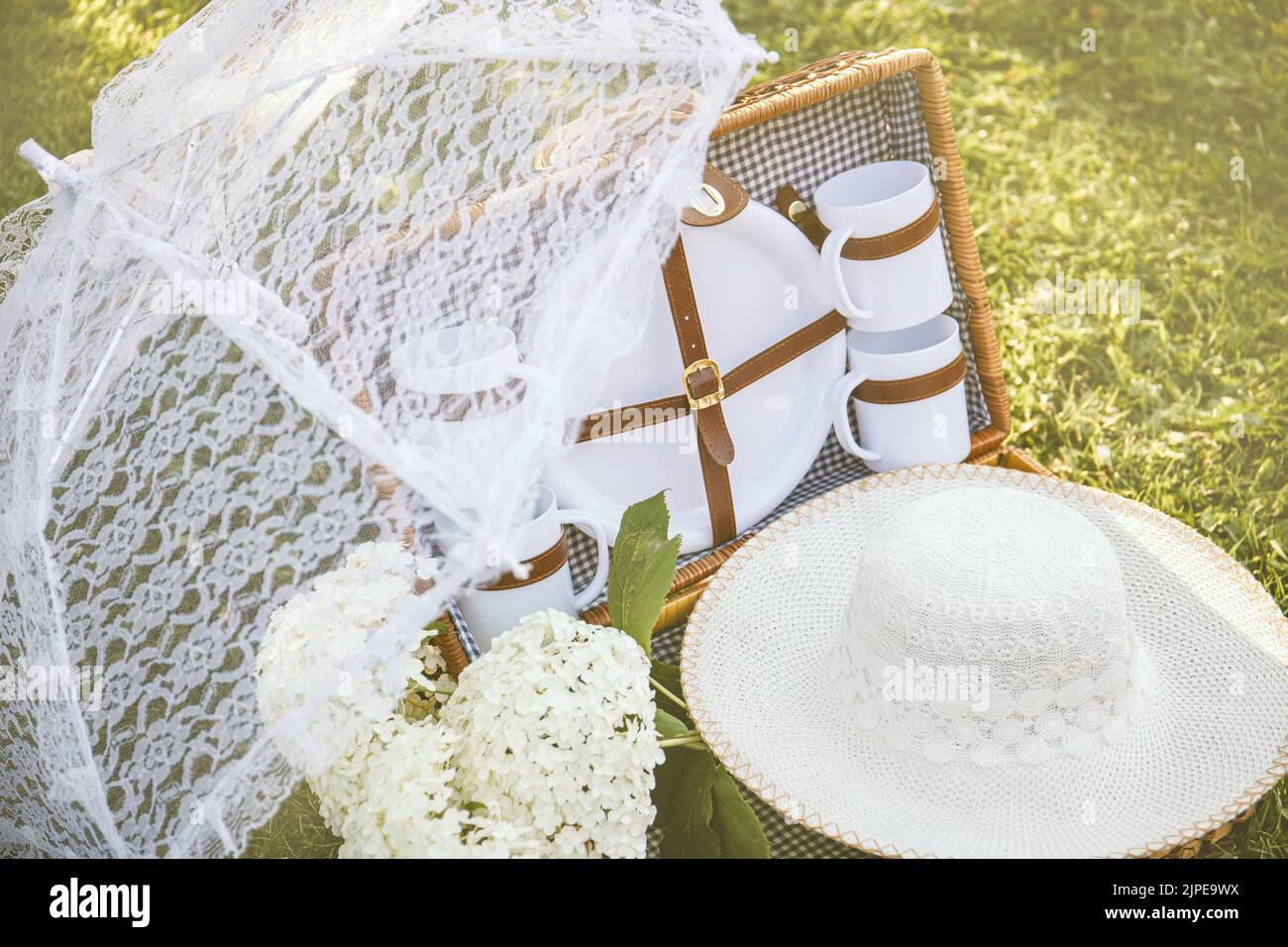 Romantisches Set mit Vintage-Picknickkorb, Spitzensonnenschirm und Sommerhut auf der Gartenrasen des Parks an sonnigen Sommertagen im Freien. Verträumt warmes Foto. Stockfoto