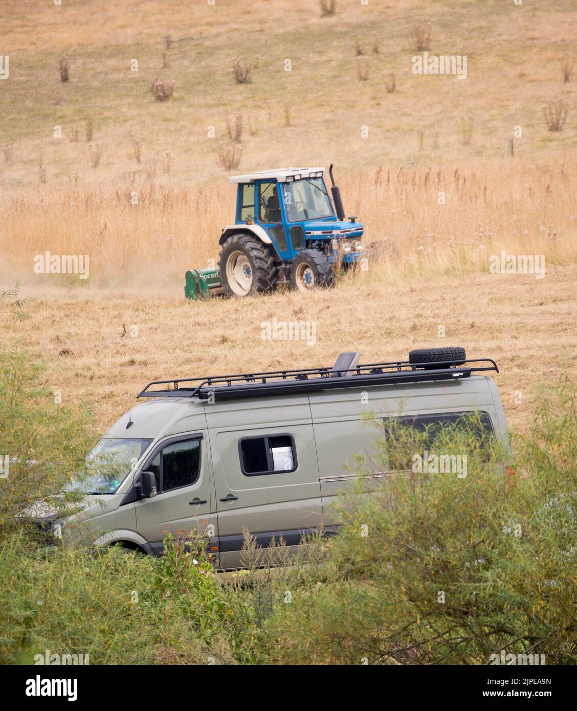 Wohnmobil parkt auf dem Land Großbritanniens, während ein Landwirt in seinem Traktor auf einem nahegelegenen Feld arbeitet. Britische Urlauber genießen Sommermobil. Stockfoto