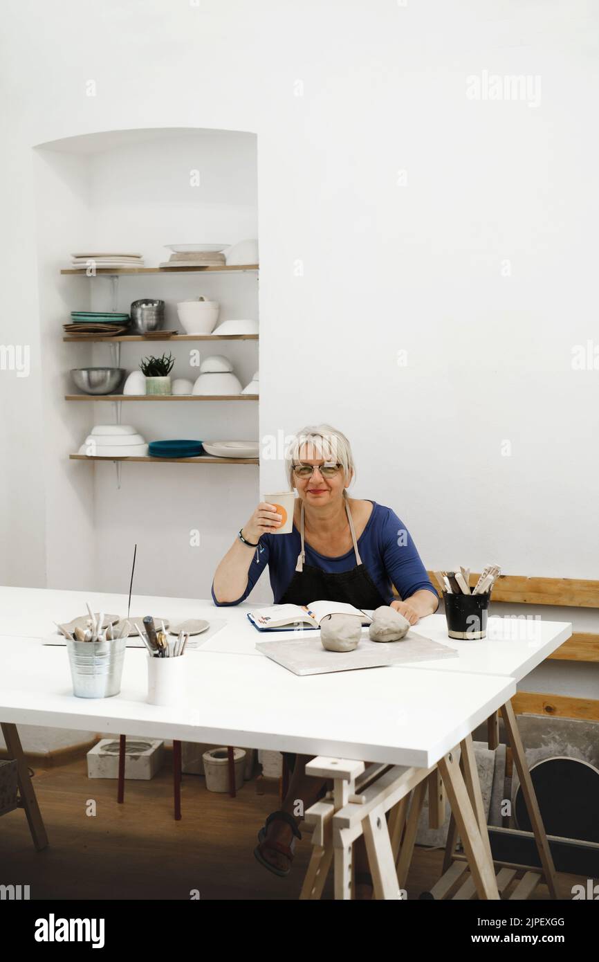 Frau Töpferin plant Arbeit in der Werkstatt, Schreiben in Notebook im Lichtstudio ihre Kreationen. Tonstücke auf dem Tisch. Kleines kreatives Unternehmen. Kopieren Stockfoto