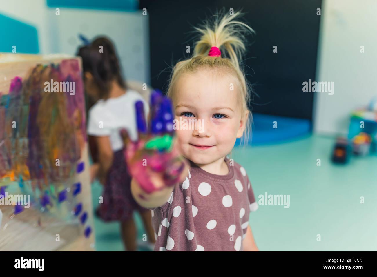 Frischhaltefolie Malerei im Kindergarten. Glücklich lächelnd blonde Kleinkind Mädchen zeigt ihre bemalte bunte Handfläche. Kunstunterricht mit Fingermalerei für Kinder sensorische Fähigkeiten, Kreativität und Vorstellungskraft Entwicklung. Hochwertige Fotos Stockfoto