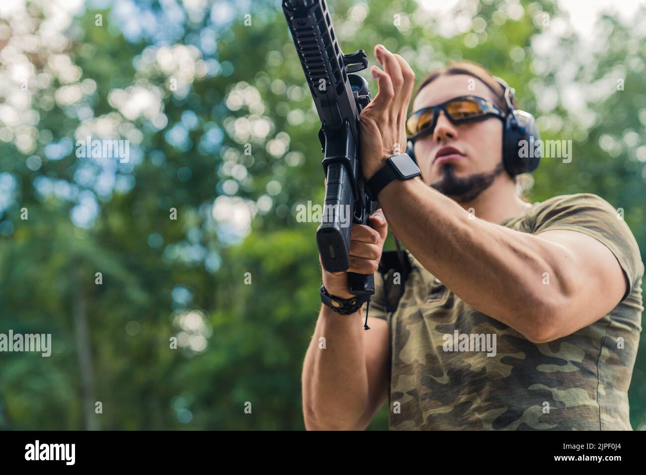 Kaukasischer bärtiger Mann mit Schutzbrille und Kopfhörern mit Camo-T-Shirt zum Nachladen der Maschinenpistole. Horizontale Aufnahme im Freien. Hochwertige Fotos Stockfoto