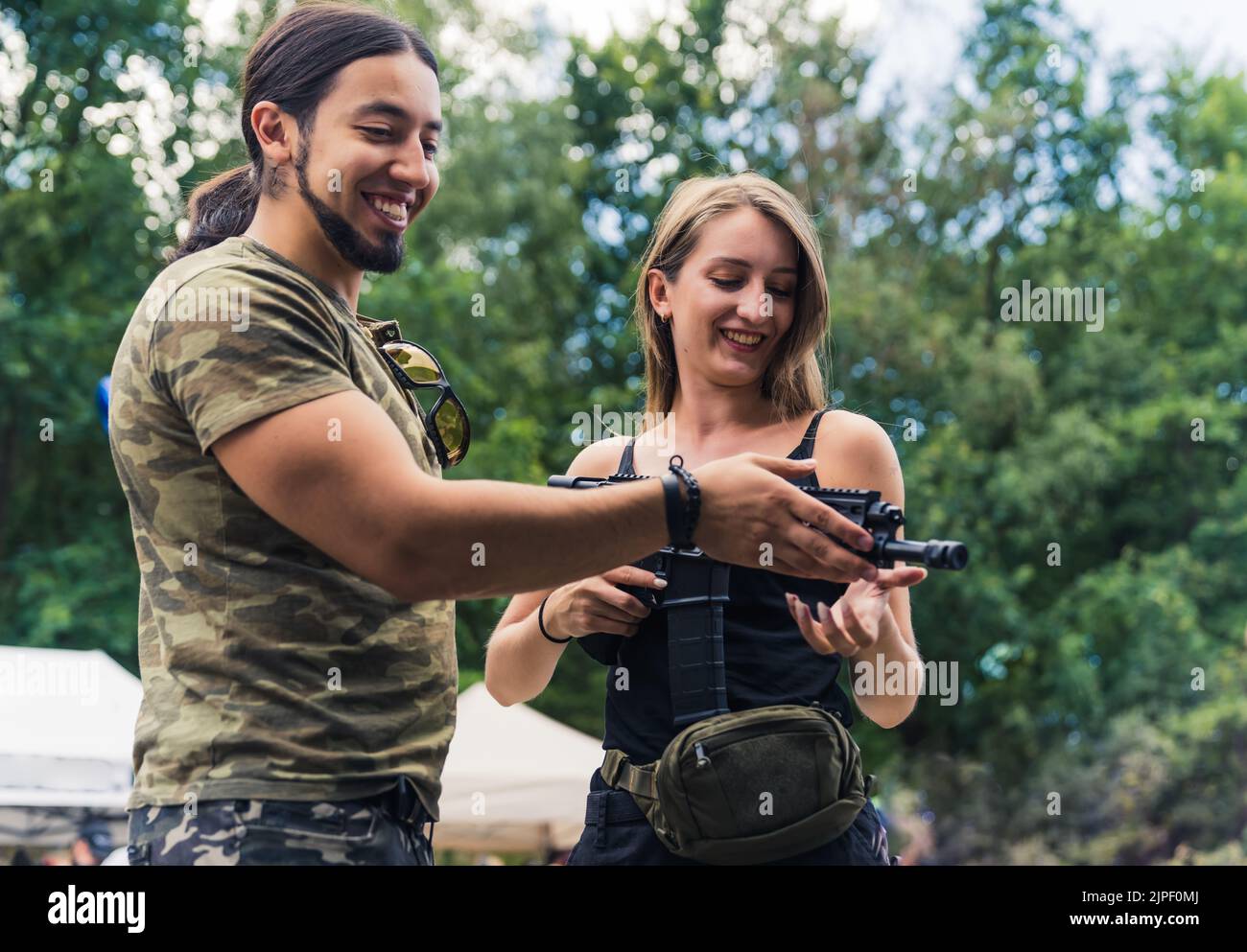 Bärtiger Mann im Camo-T-Shirt mit Maschinengewehr für weibliche Klientin. Kauf und Verkauf von Schusswaffen Thema. Horizontale Aufnahme im Freien. Hochwertige Fotos Stockfoto