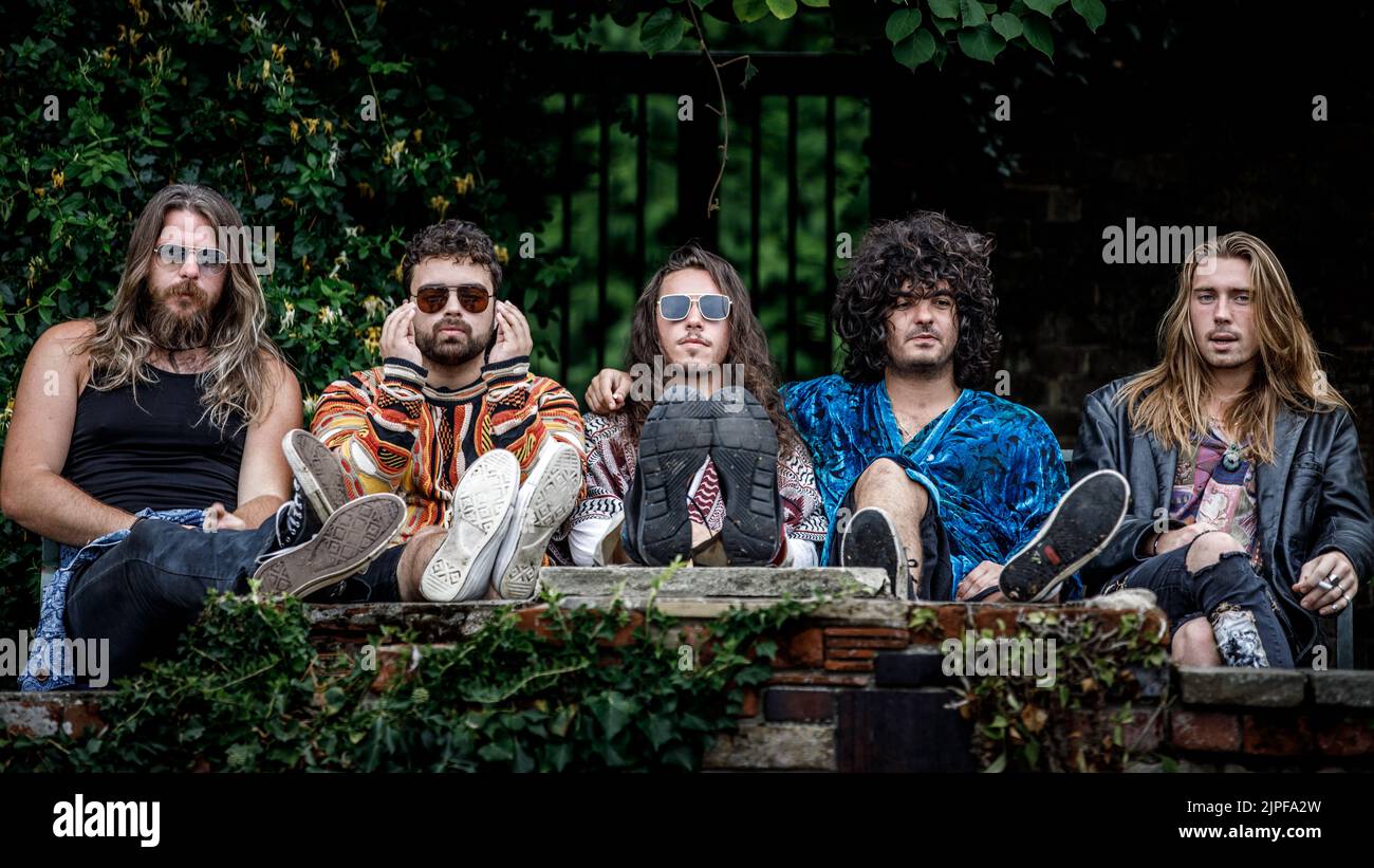 Fünf junge Männer in coolen Kleidern sitzen auf einer Bank in einem Garten. Rockgruppe, Musiker, Langhaarmänner zusammen Stockfoto