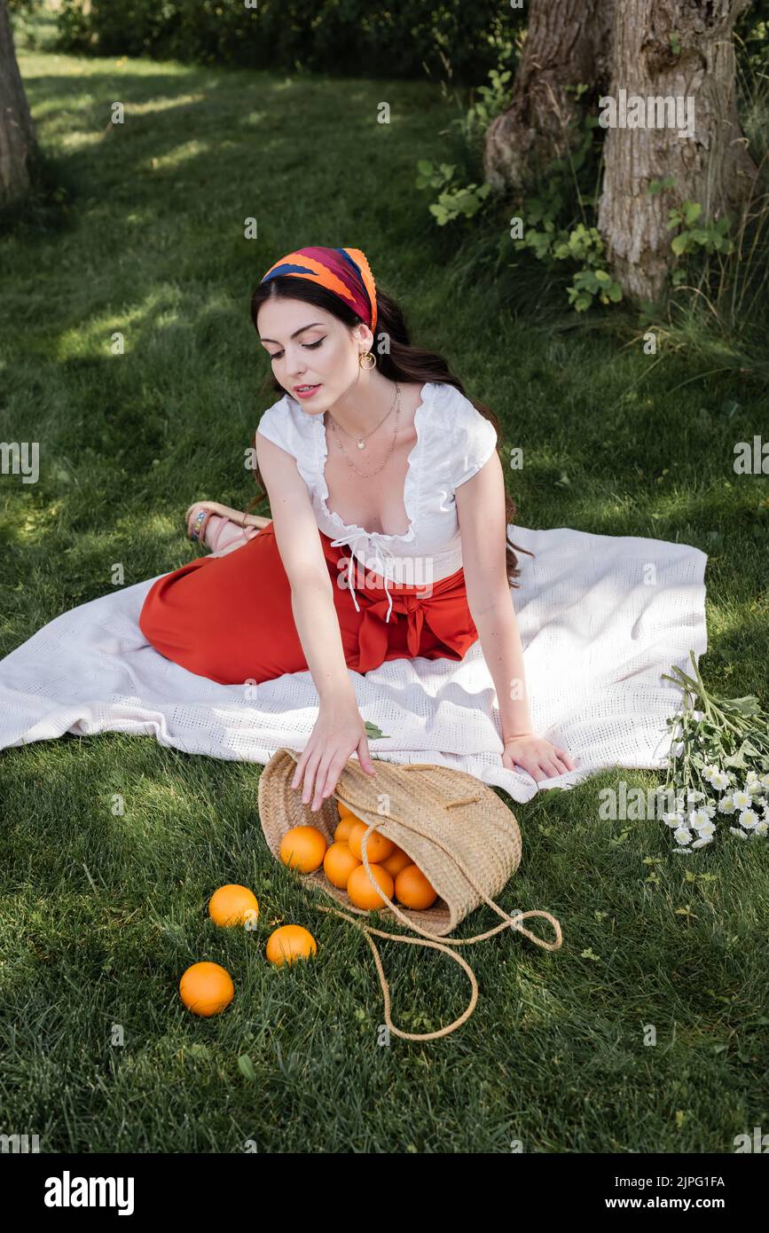 Junge modische Frau, die Orangen in der Tasche in der Nähe von Blumen im Park betrachtet Stockfoto